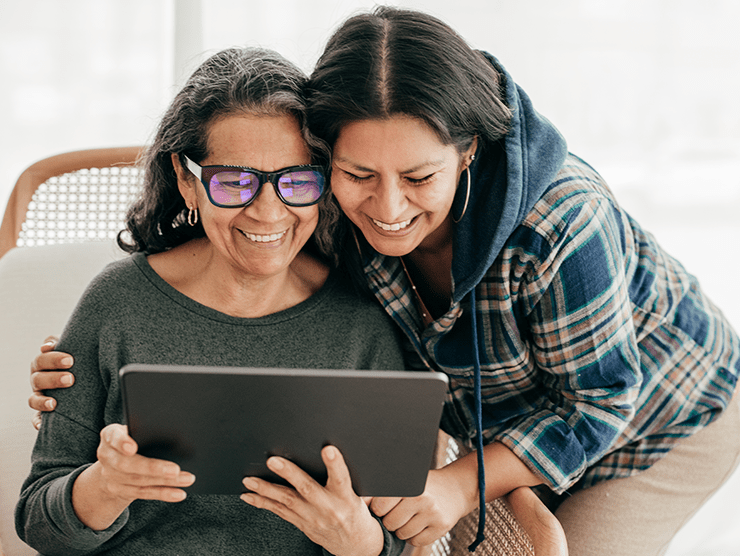 A woman affectionately holding her mom while they are both looking at a tablet smiling.