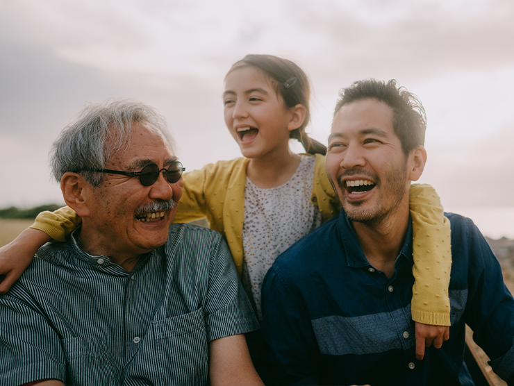 An elderly man with his son and granddaughter smiling outside.