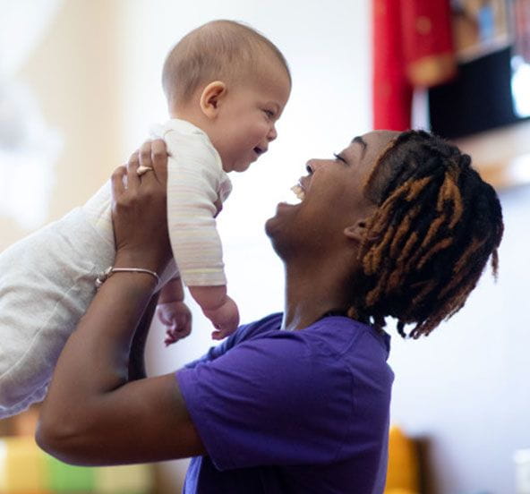 A teacher holds an infant in the air at daycare