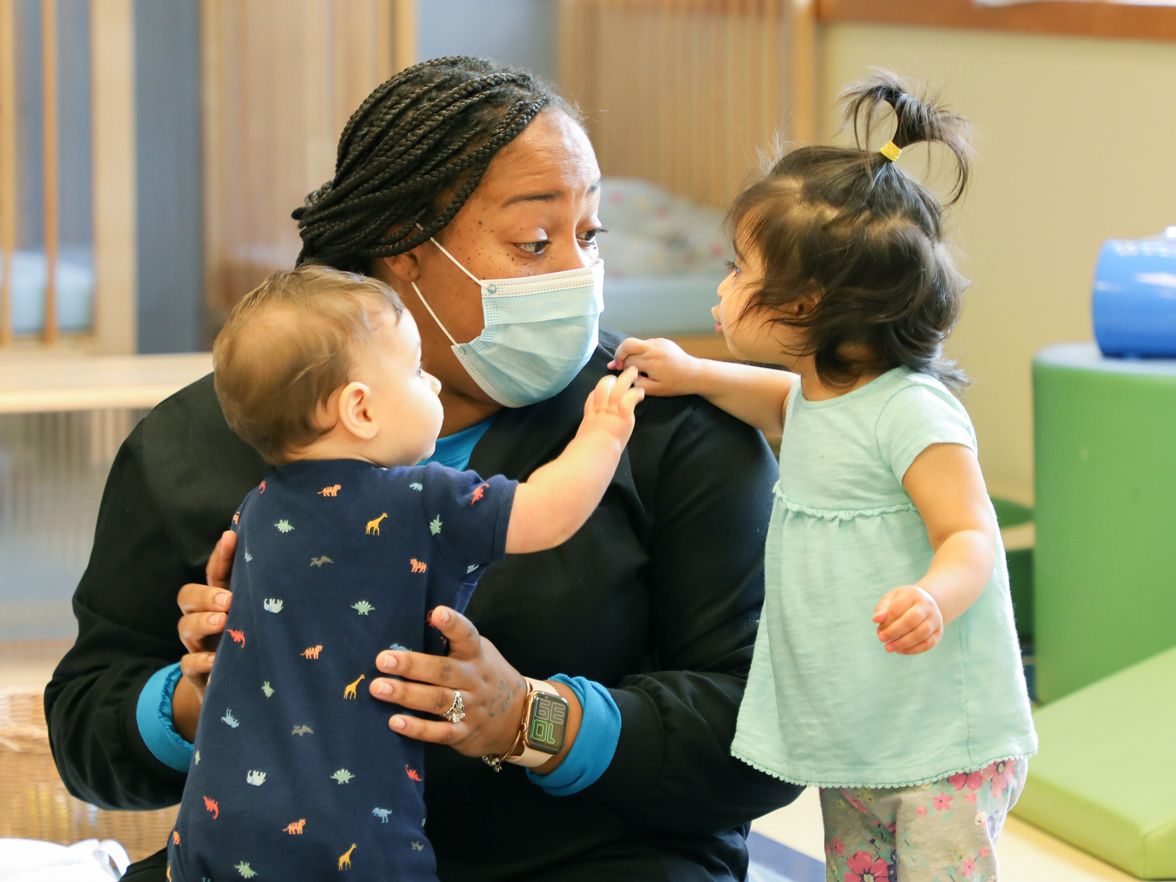 Teacher practicing safe health and safety wearing a mask, in front of two young children | Bright Horizons | Daycare