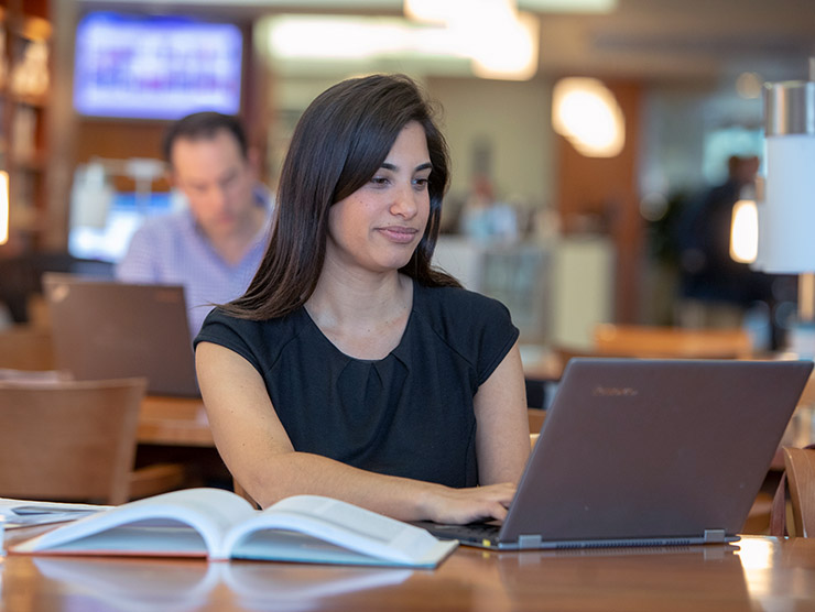 Woman in library working on laptop with textbook open