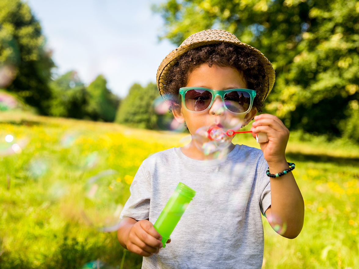 Young boy blows bubbles outside with hat and glasses on at a Bright Horizons Summer Camp Daycare program