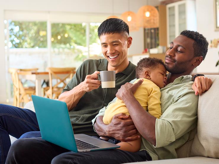 Two dads sitting on couch looking at a laptop with a sleeping baby