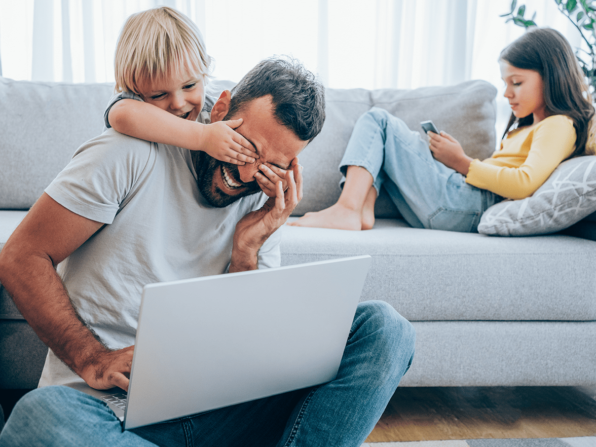 Father working on a laptop with child covering his eyes