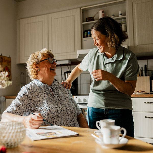 Older woman sitting at a kitchen table with a magazine in front of her, smiling at a younger woman standing next to her
