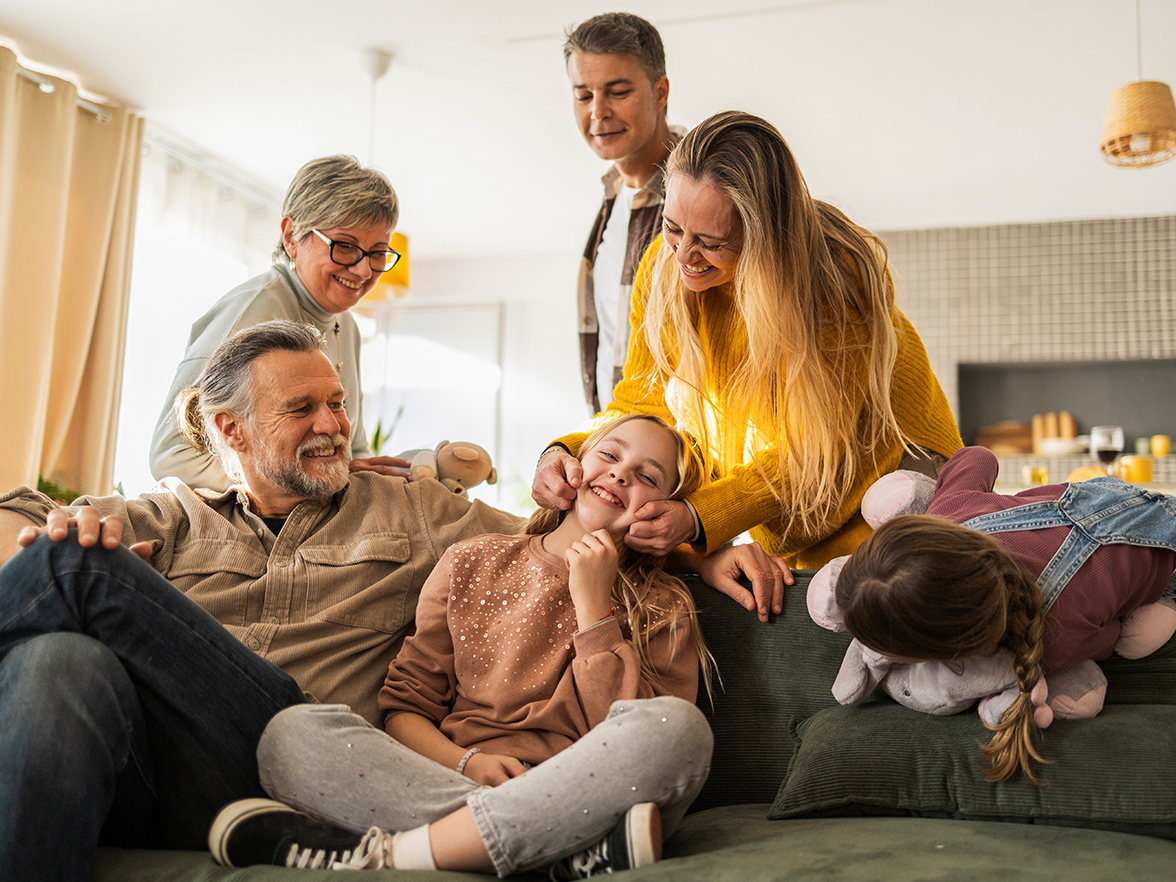 Grandpa sitting with two grandkids on the couch while mom, dad, and grandma stand behind them — all smiling