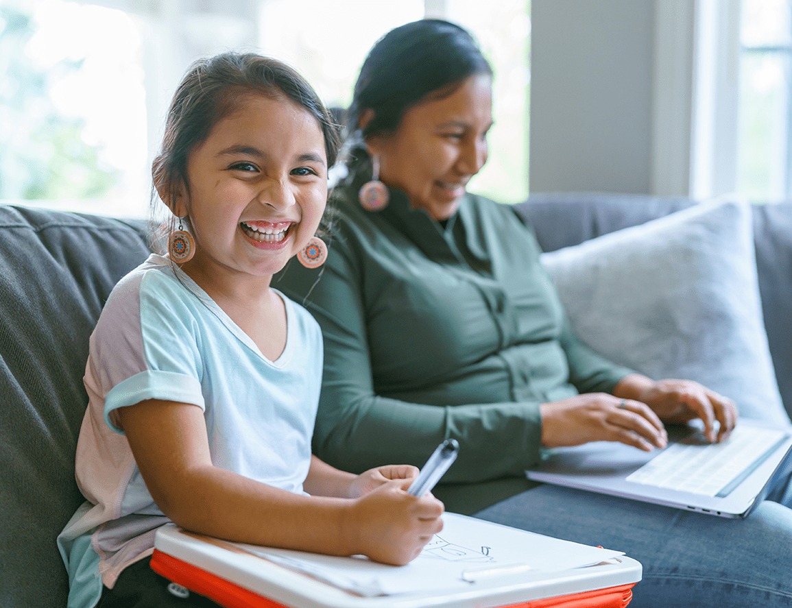 Child doing homework with pen and notebook beside an adult using a laptop on a sunlit living room couch. 
