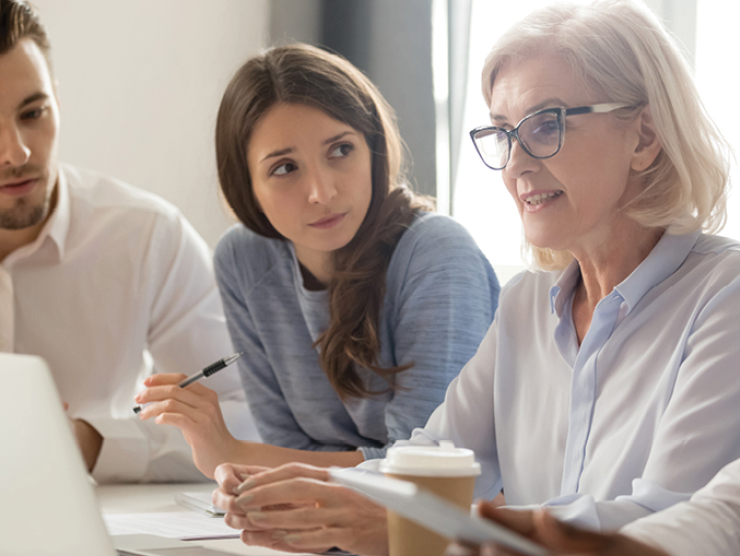 A group of people sitting together at a table in a meeting, discussing documents and working on a laptop with a coffee cup in the foreground.
