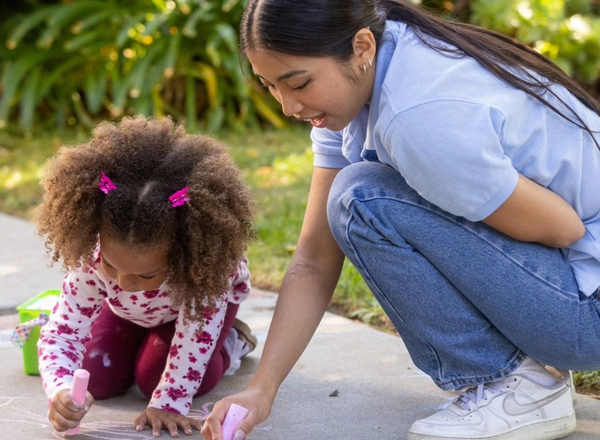Two girls, a child and teacher, draw with chalk on a sidewalk. 