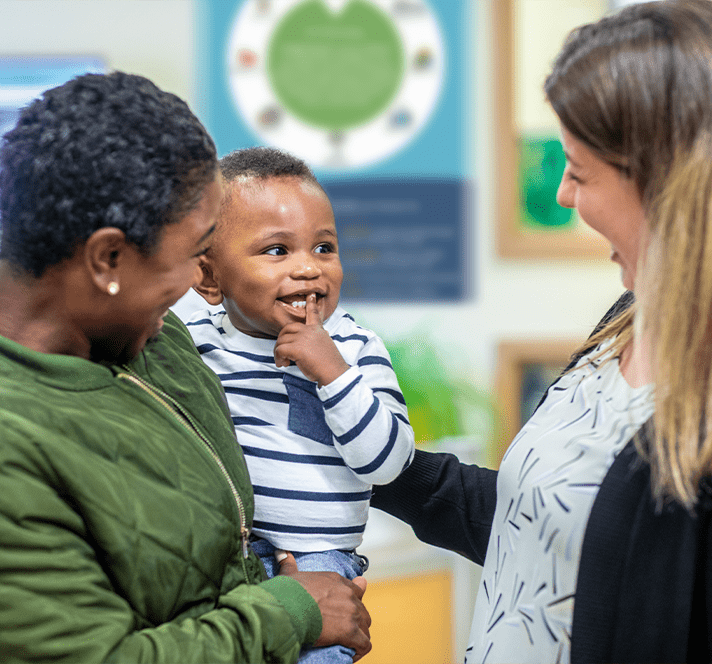 A mother holds her smiling baby near a teacher. 