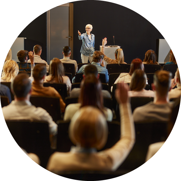 A presenter stands at the front of a dark-walled room while a seated audience faces the presentation area. 