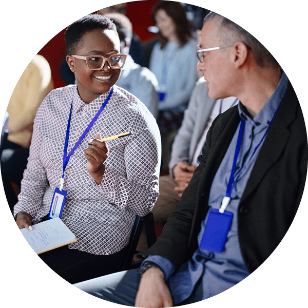 Two seated conference attendees converse in the foreground, with additional people blurred behind them. 