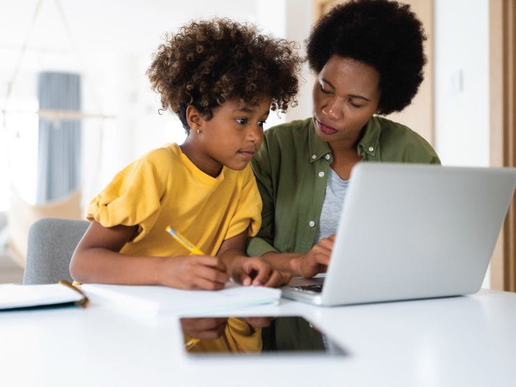 A school-aged child holding a pencil while looking at a laptop screen with an adult next to her.