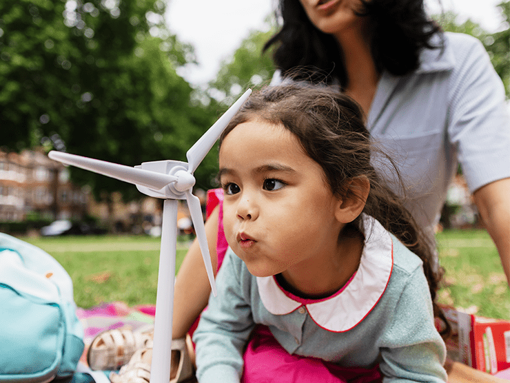 A young girl sitting at the park with her mom blowing on a wind spinner.