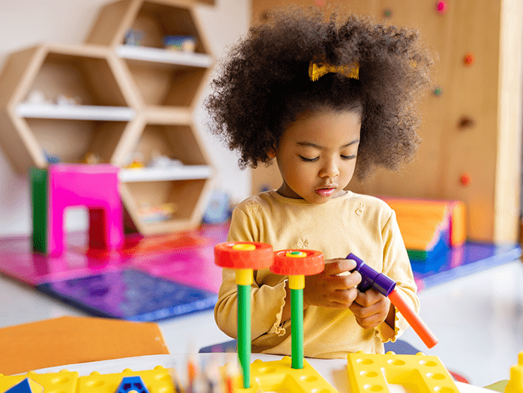 A preschool girl sitting at a table in the classroom, building a structure with colorful tubes. 