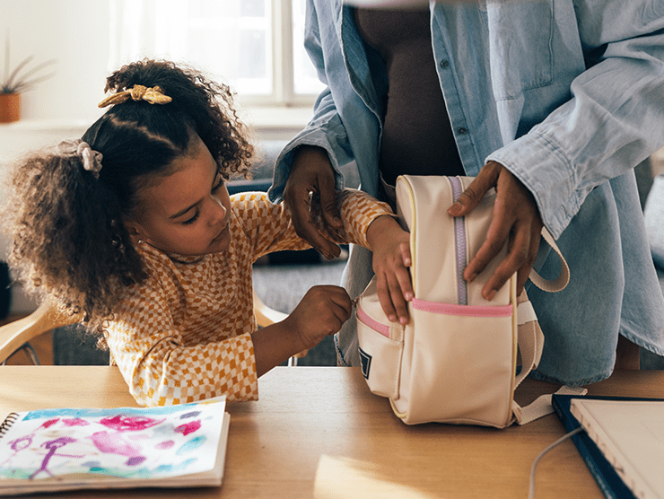 A preschool child packing her backpack while parent helps