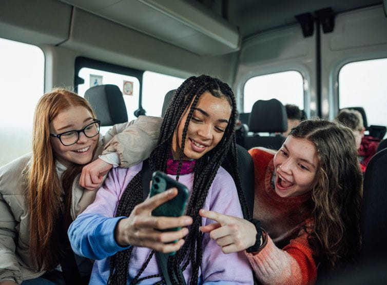 Three teenage girls sitting on a school bus laughing at something on a phone.