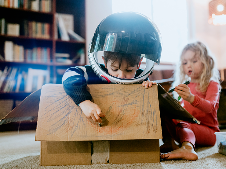 A young boy with a helmet on drawing on a cardboard box at home.