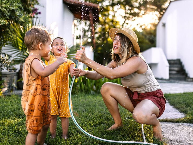 A woman and two toddler children playing with water from a hose outdoors