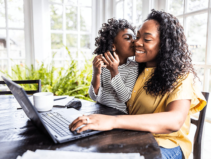 A child sitting on his mom's lap giving her a kiss on the cheek, while the mom is on her laptop