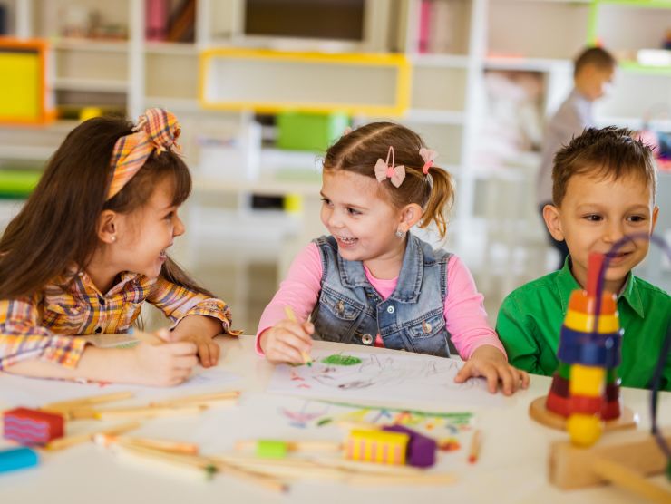 Two preschool aged children in a classroom setting sitting at a table drawing while smiling at each other.