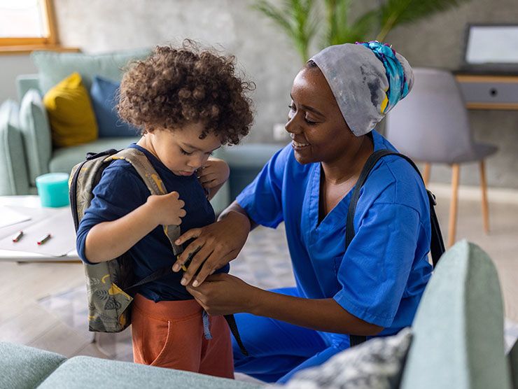 A mom helping her young child put on his backpack.