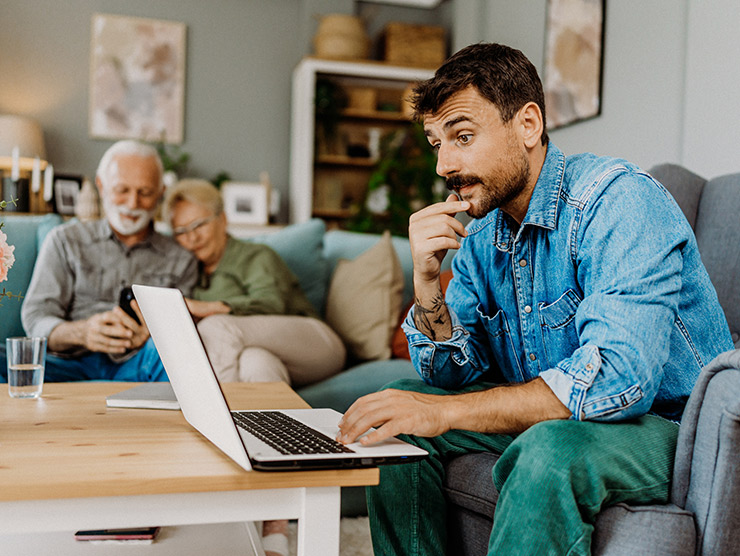 Adult male working on laptop on couch while elderly parents are sitting nearby on their phone.