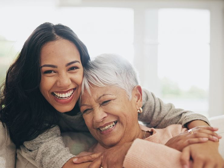 Young woman smiling at the camera, with her arms around her older mother.