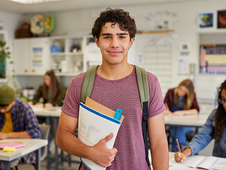 A teenage male with a backpack on, holding books and smiling at the camera in a classroom setting