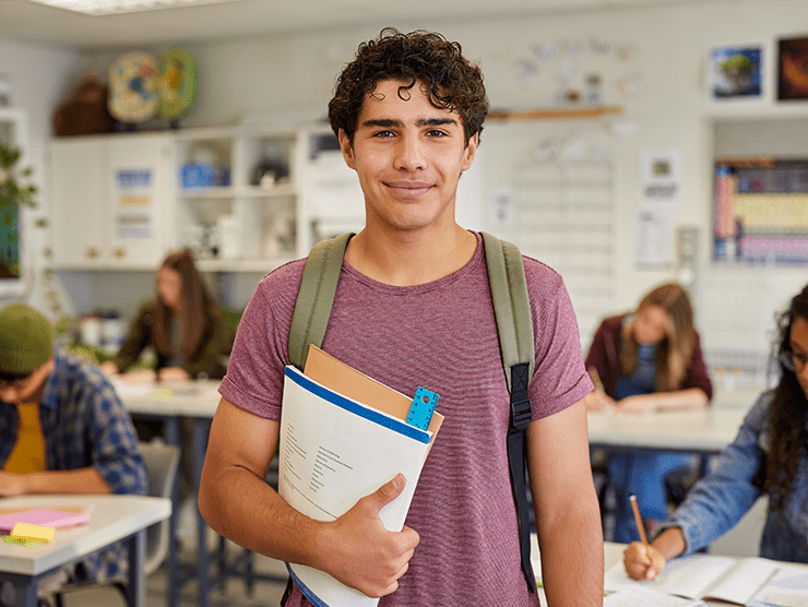 A teenage male with a backpack on, holding books and smiling at the camera in a classroom setting