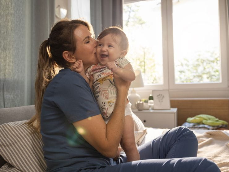 A nurse in scrubs sitting at home kissing her baby.