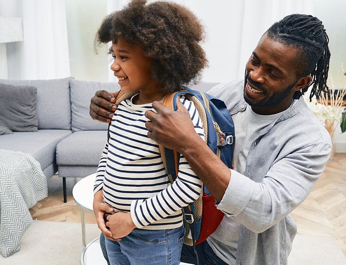 A dad helps daughter getting ready to school and puts a school bag on her back.