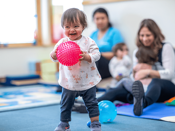 A toddler child standing while holding a ball in a playroom setting.