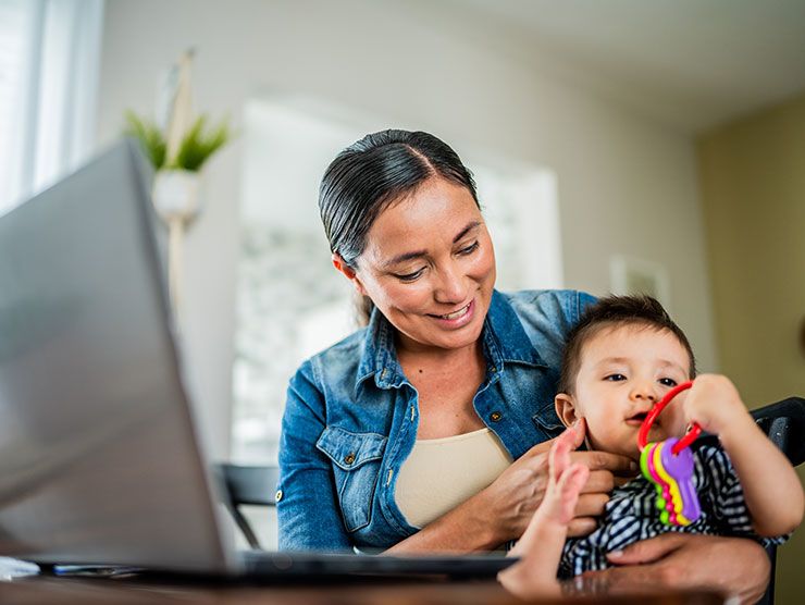 A mom holding her baby while sitting at her computer.