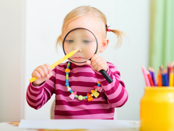A young girl using a magnifying glass to observe a yellow pencil in a classroom setting