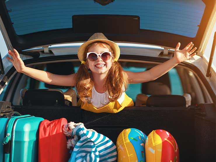 A girl in a hat and sunglasses looking out the back of a car smiling at the camera