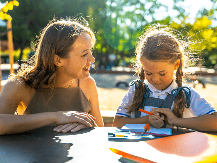 A young child sitting with an adult woman in a park with markers in her hand.