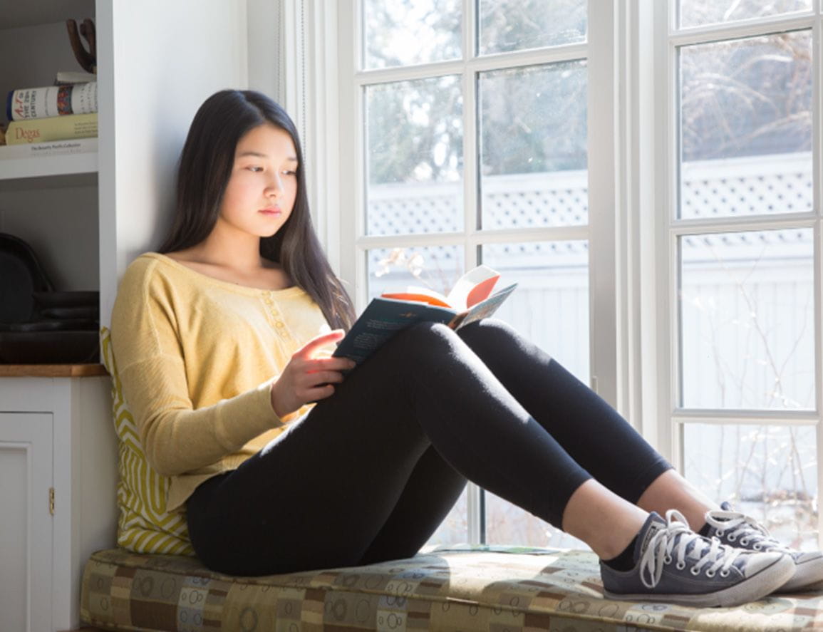 A teenage girl reading by her window.
