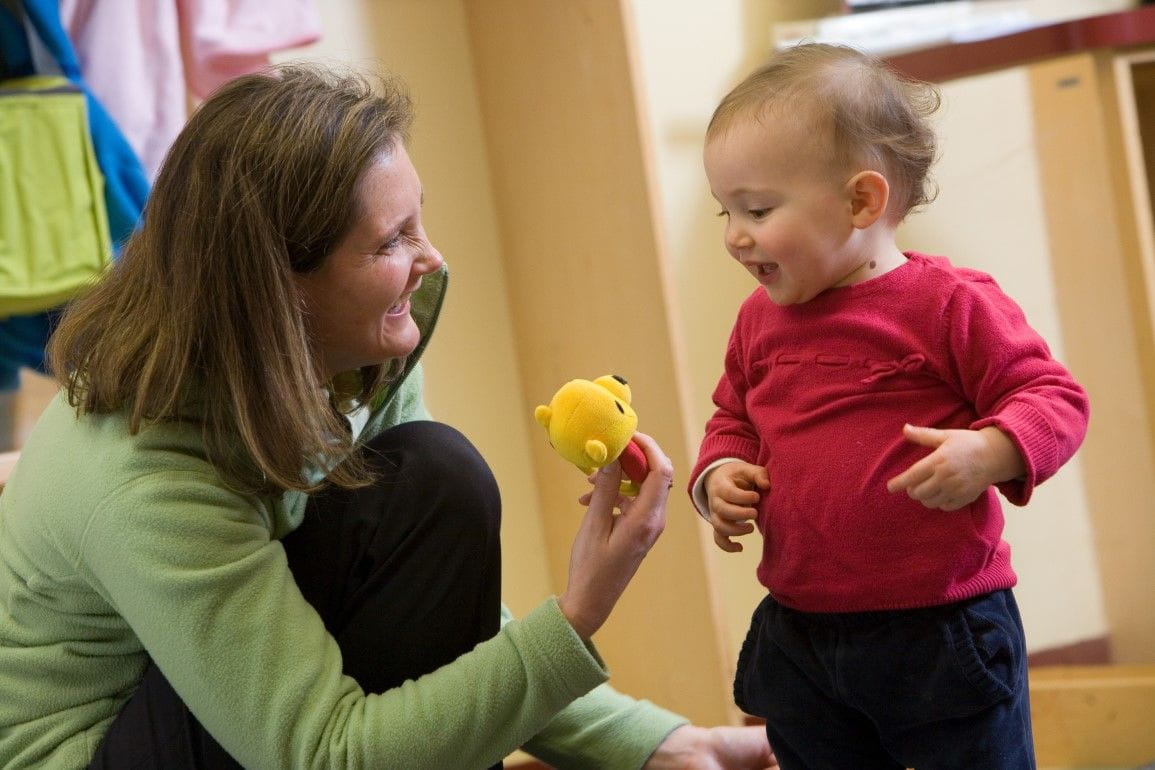 Woman kneels down to give a child a yellow toy at daycare