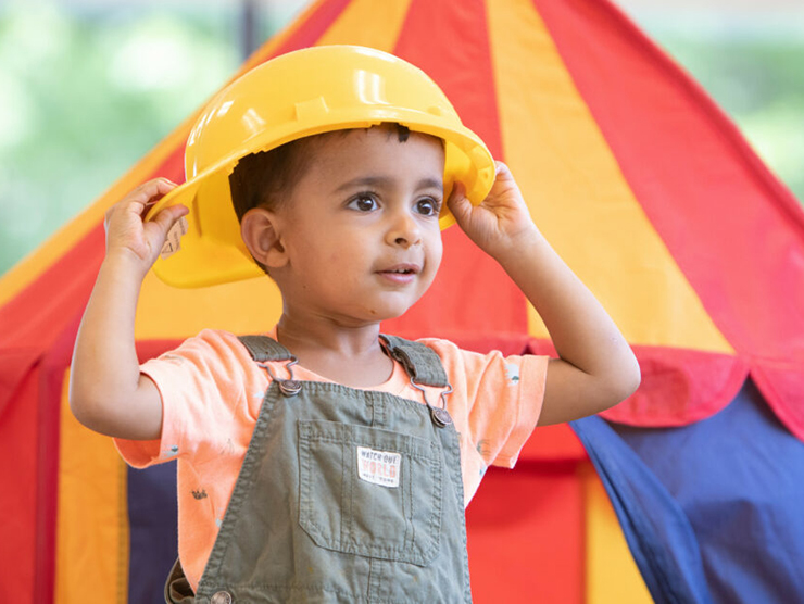 Toddler playing with a construction hat while outside in front of a tent 