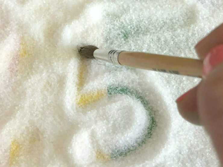 Preschooler using a paintbrush to write numbers on a tray of salt 