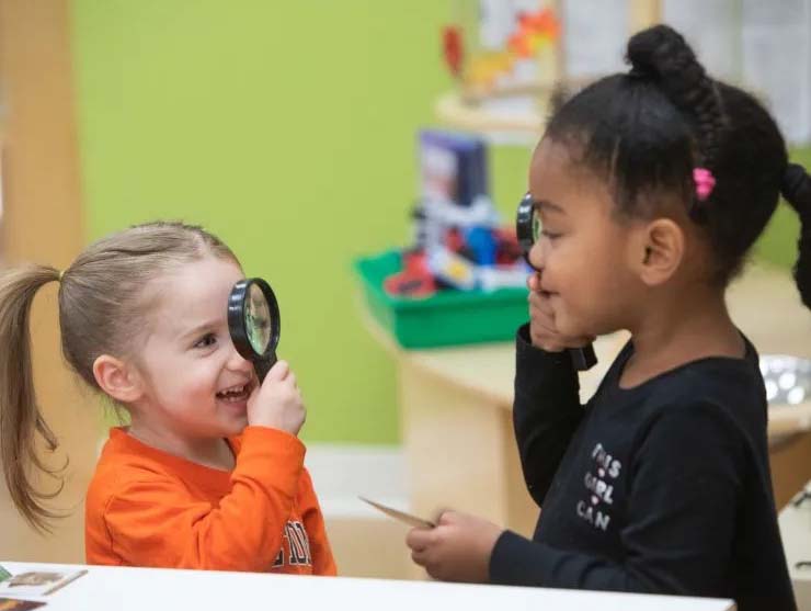 Two children smile and laugh as they look through magnifying glasses | Bright Horizons