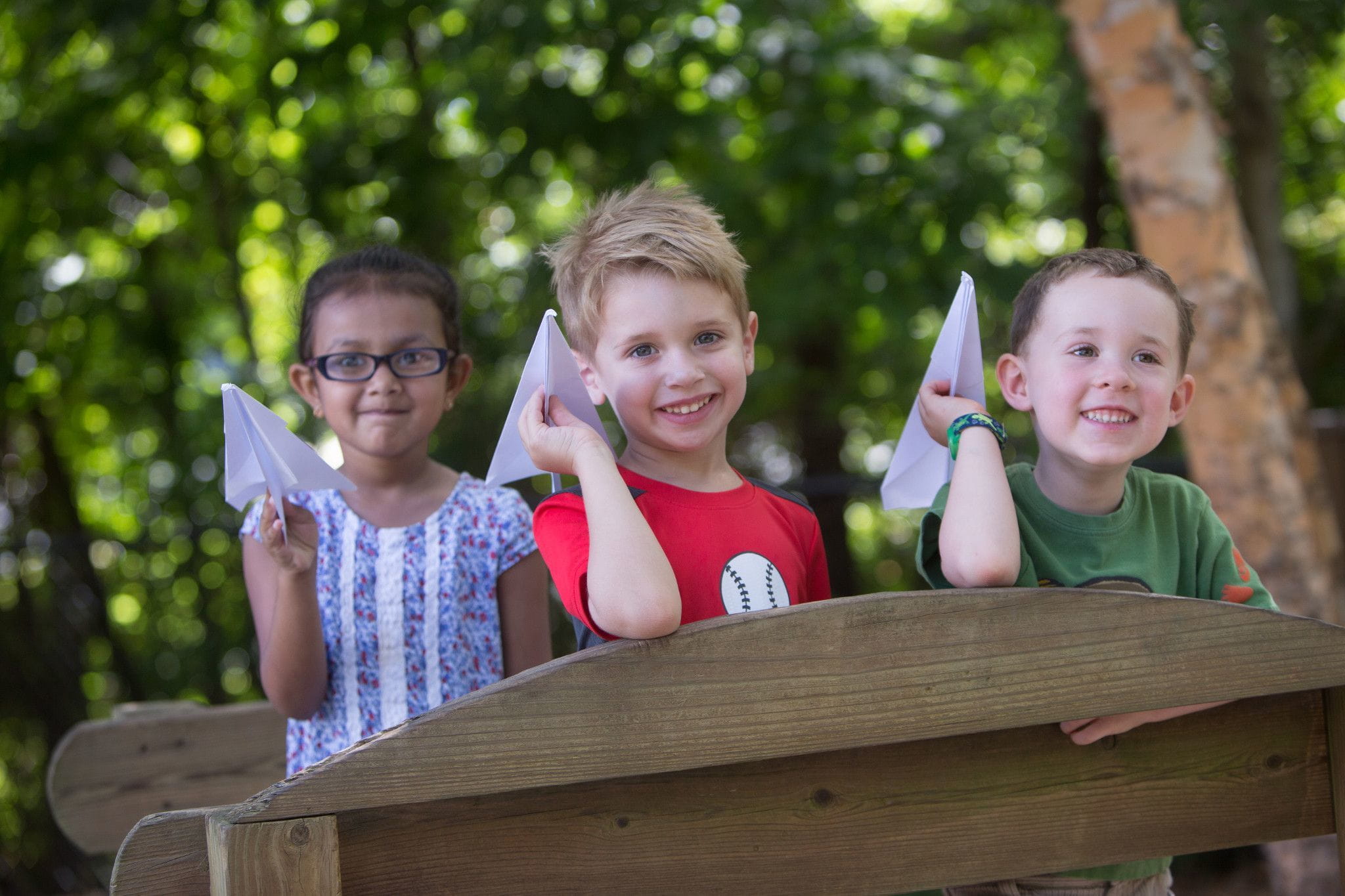 3 children smiling and playing outside with paper airplanes